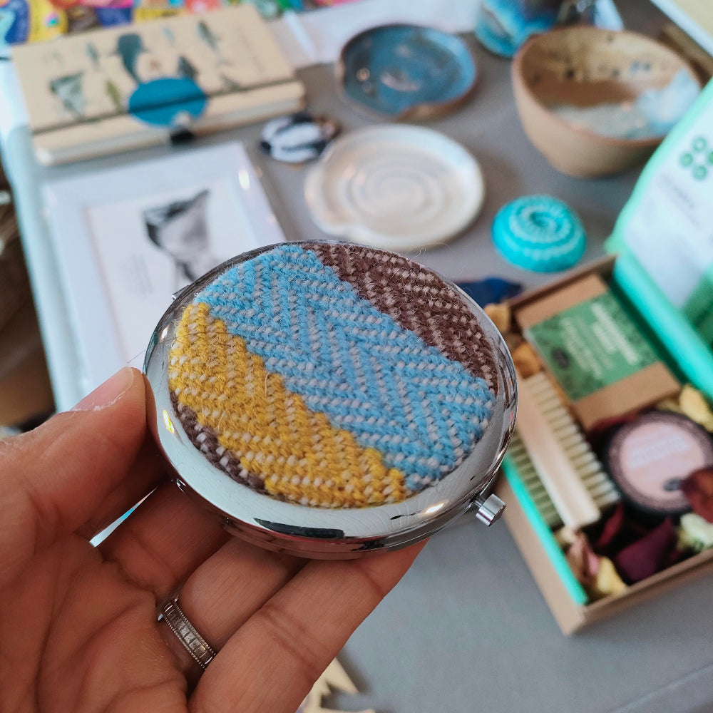 Compact mirror with textured blue and yellow design held in a hand, with a blurred background.