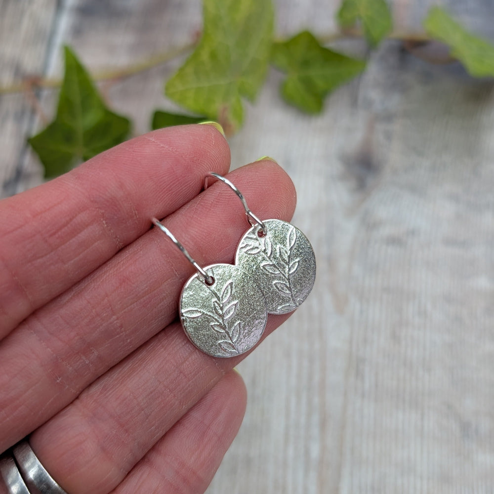 A pair of silver leaf-shaped dangle earrings held on a person's fingers, with blurred green leaves and a light wood surface in the background.