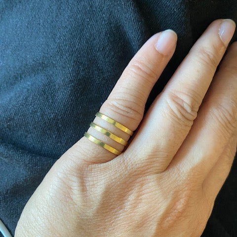 A close-up of a person's hand wearing three thin gold bands stacked together on one finger, resting against a dark charcoal-colored fabric background.
