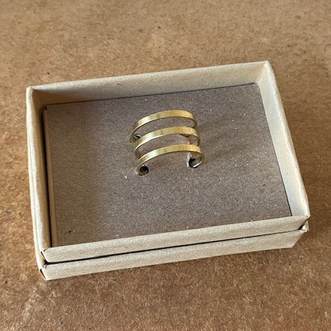 A gold-toned three-band ring displayed inside an open beige cardboard jewelry box on a brown textured surface.