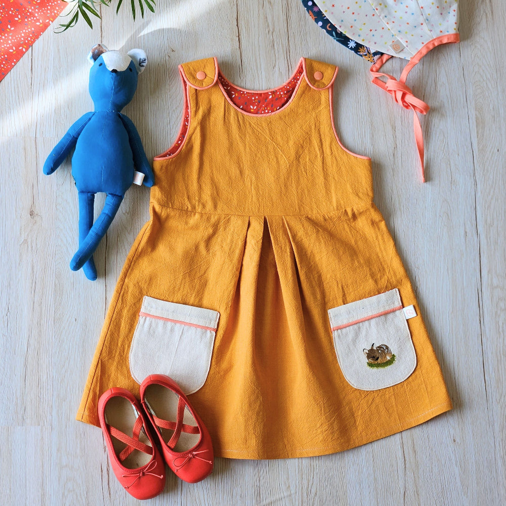 A baby girl's mustard yellow linen pinafore dress with white front patch pockets, laid flat on a light wood floor alongside a blue plush toy, a white polka-dot sun hat, and red ballet flats.