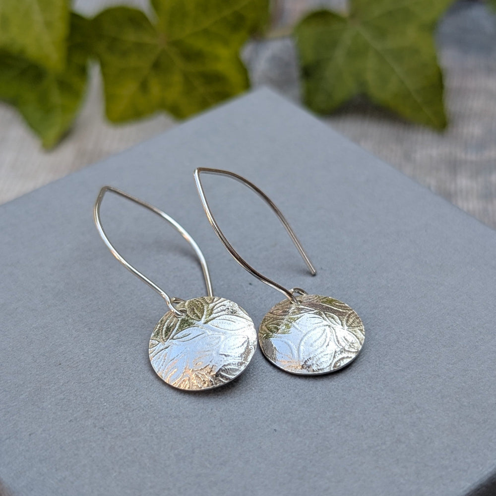 A pair of silver drop earrings with textured, hammered circular discs and curved wire hooks, displayed on a gray square block with blurred green leaves in the background.